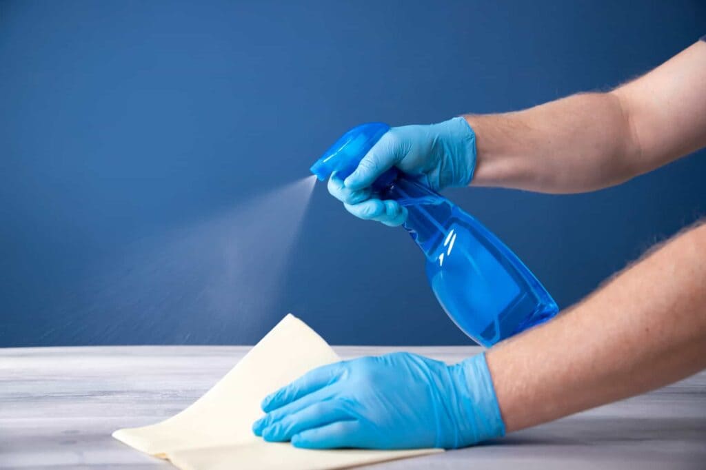 Person wearing gloves disinfecting a surface with a blue antiseptic spray bottle during the coronavirus pandemic.